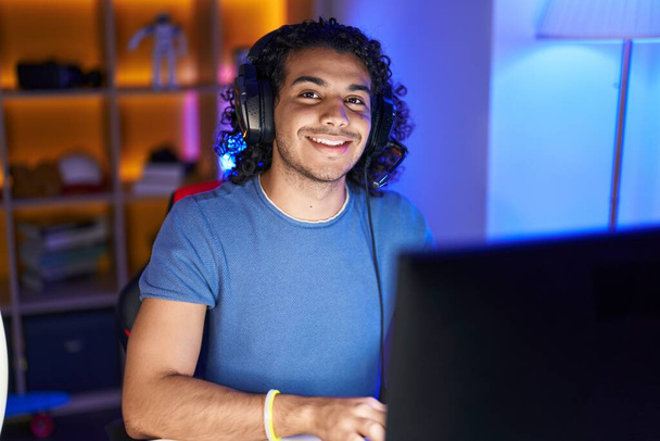 jong latin man streamer glimlachen zelfverzekerd zitten op tafel bij gaming kamer - Foto, afbeelding