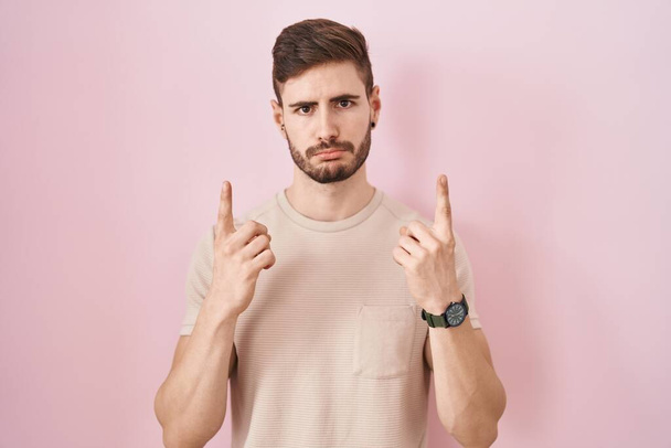 Uomo ispanico con la barba in piedi su sfondo rosa che punta verso l'alto guardando triste e sconvolto, indicando la direzione con le dita, infelice e depresso.  - Foto, immagini