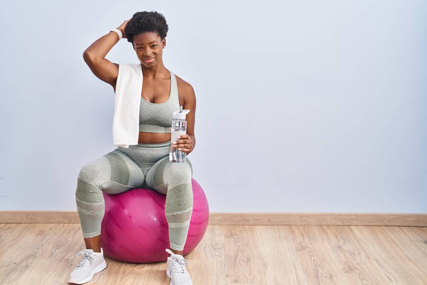 African american woman wearing sportswear sitting on pilates ball confuse and wondering about question. uncertain with doubt, thinking with hand on head. pensive concept.  - Fotó, kép