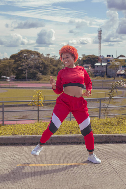 Joven afro de cabello naranja bailando al estilo urbano en parque local con pista de atletismo, vegetacion y cielo hermosos de fondo - Φωτογραφία, εικόνα