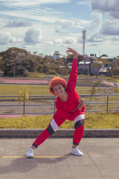 Joven afro de cabello naranja bailando al estilo urbano en parque local con pista de atletismo, vegetacion y cielo hermosos de fondo - Foto, immagini