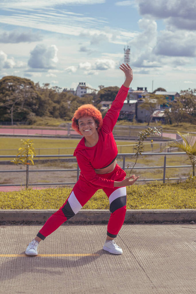 Joven afro de cabello naranja bailando al estilo urbano en parque local con pista de atletismo, vegetacion y cielo hermosos de fondo - Foto, imagen