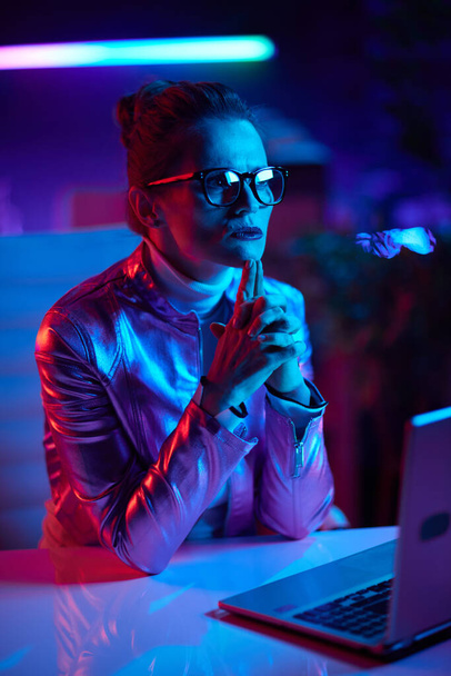 Neon metaverse futuristic concept. pensive trendy 40 years old woman in glasses with laptop in modern office. - Foto, Bild