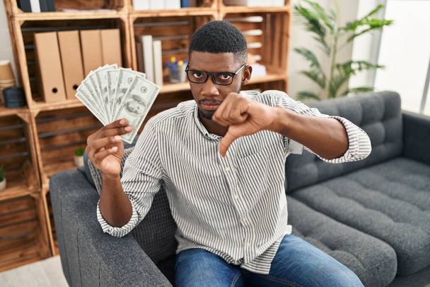 Afrikai amerikai férfi kezében dollár dühös arc, negatív jel mutatja, nem tetszik hüvelykujjal lefelé, elutasítás koncepció  - Fotó, kép