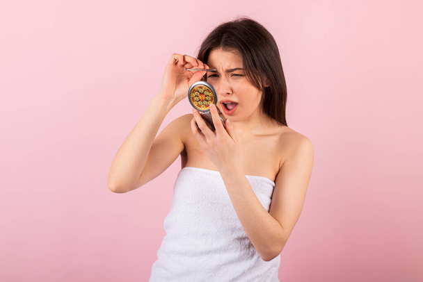attractive teenage girl wrapped only in a white towel is looking at herself in a small portable mirror and plucking her eyebrows with tweezers. isolated on a pink background. - Photo, image