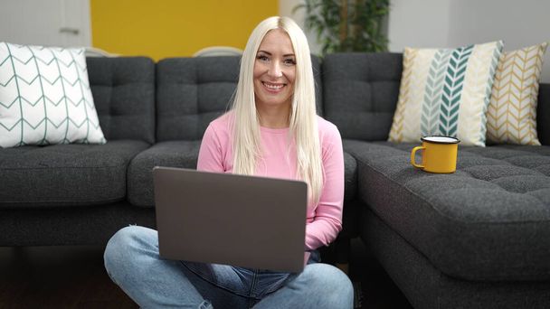 Молода блондинка використовує ноутбук, сидячи на підлозі вдома
 - Фото, зображення