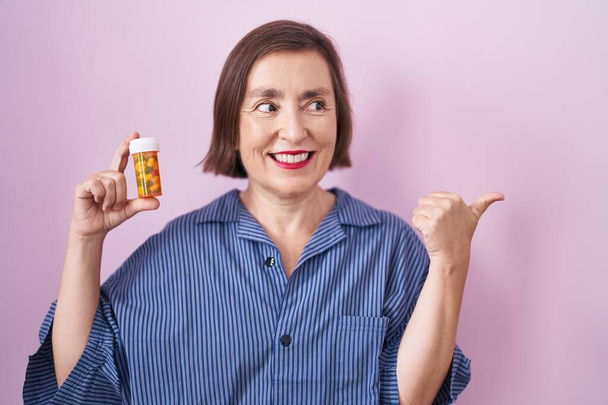 Middle age hispanic woman holding pills pointing thumb up to the side smiling happy with open mouth  - Foto, imagen