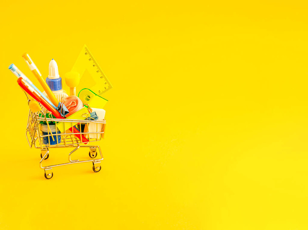 Stationery accessories in a shopping trolley on a yellow background with place for text. School preparation concept, flat lay, copy space. View from above. - Фото, изображение
