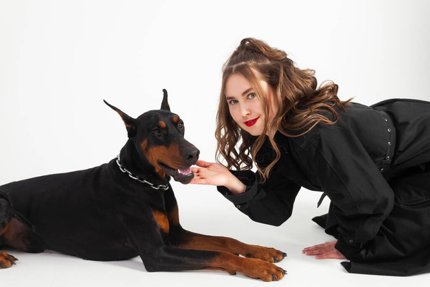 A young beautiful Doberman with a chain around his neck and his owner, elegant girl in a long black coat and bright red lipstick. Isolated on a white background. - Фото, зображення