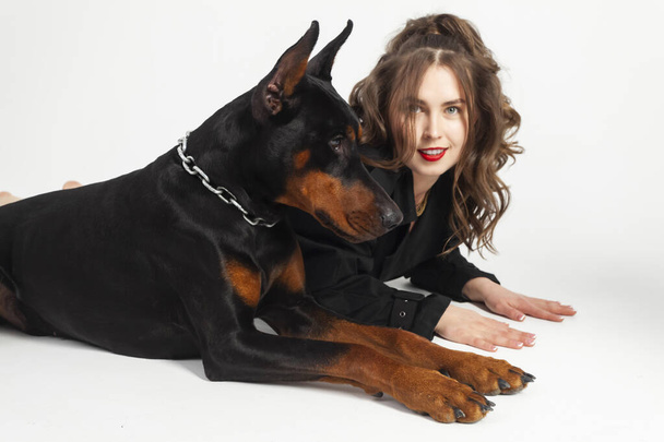 A young beautiful Doberman with a chain around his neck and his owner, elegant girl in a long black coat and bright red lipstick. Isolated on a white background. - Foto, afbeelding