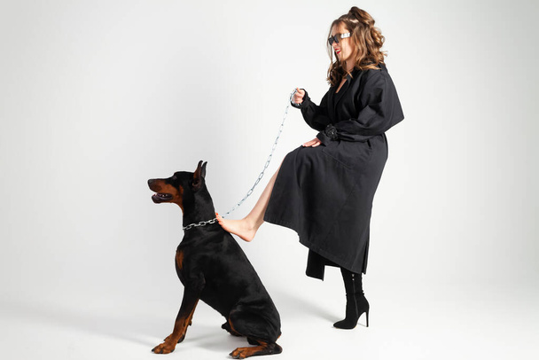 A young beautiful Doberman on a chain and his owner, elegant girl in a long black coat and bright red lipstick, lost one shoe. Isolated on a white background. - Photo, Image