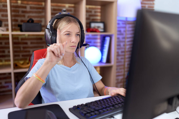 Mujer caucásica joven jugando videojuegos con auriculares apuntando con el dedo hacia arriba y la expresión enojada, que no muestra ningún gesto  - Foto, imagen