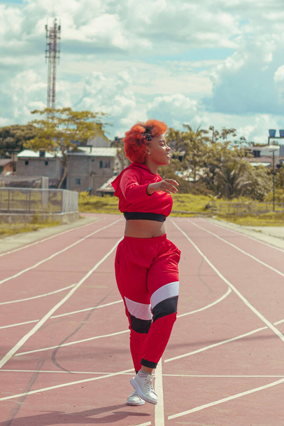 Joven afro de cabello naranja bailando al estilo urbano en parque local en la pista de atletismo, vegetacion y cielo hermosos de fondo - Photo, Image