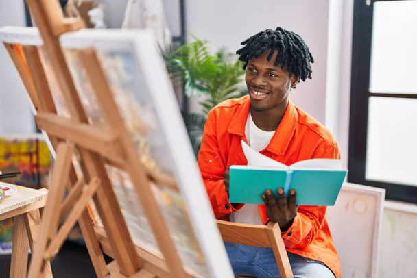 Αφροαμερικάνος καλλιτέχνης χαμογελώντας αυτοπεποίθηση ανάγνωση βιβλίο σχέδιο στο στούντιο τέχνης - Φωτογραφία, εικόνα
