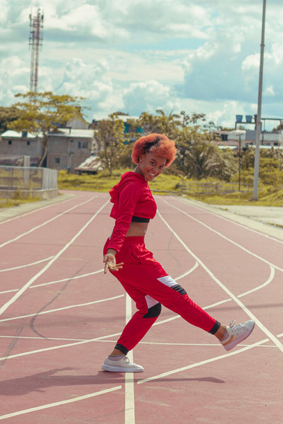 Joven afro de cabello naranja bailando al estilo urbano en parque local en la pista de atletismo, vegetacion y cielo hermosos de fondo - 写真・画像