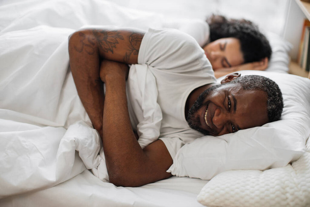 Smiling bearded husband relaxing on bed with pillow under head while sleeping wife resting on background. Cheerful african american man in mature relationship relishing full life. - Fotó, kép