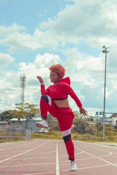 Joven afro de cabello naranja bailando al estilo urbano en parque local en la pista de atletismo, vegetacion y cielo hermosos de fondo - Foto, Imagen