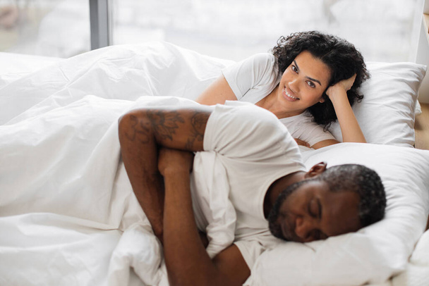 Pretty african american woman leaning on soft pillow already awake while her attractive husband still getting shuteye on bed in front of panoramic window. Happy couple spending morning together. - Фото, изображение