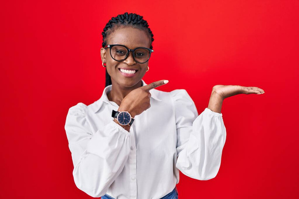 Afrikanerin mit Zöpfen steht erstaunt vor rotem Hintergrund und lächelt in die Kamera, während sie mit der Hand präsentiert und mit dem Finger zeigt.  - Foto, Bild