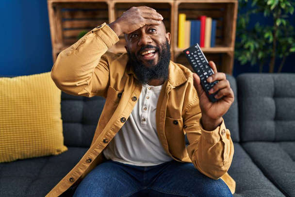 Afrikkalainen amerikkalainen mies jolla televisio kaukosäädin stressaantunut ja turhautunut käsi pään päällä, yllättynyt ja vihainen kasvot  - Valokuva, kuva