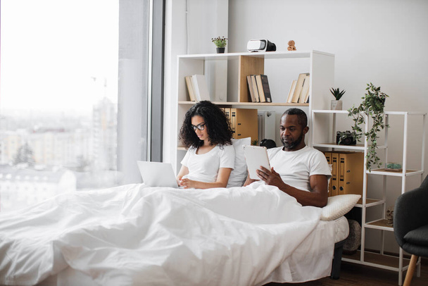 African american romantic partners using electronic devices while lounging on wide bed near panoramic window. Beautiful woman with laptop and relaxed man with tablet enjoying leisure time at home. - Fotoğraf, Görsel