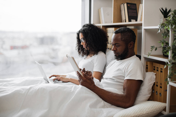 Attractive african american man and pretty woman using digital devices while leaning against soft pillows in bed of studio room. Busy spouses doing their own business without disturbing each other. - Fotoğraf, Görsel