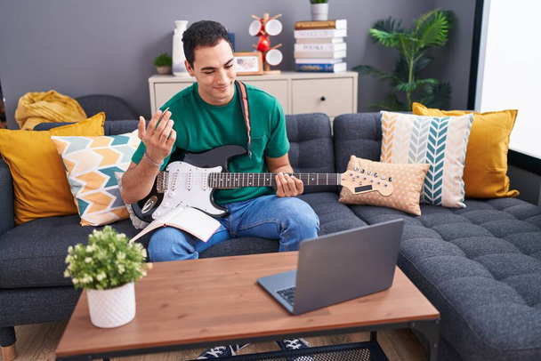 Jonge Spaanse man met online elektrische gitaar klasse zittend op de bank thuis - Foto, afbeelding