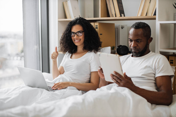 Pretty family woman keeping portable computer on blanket and showing thumb-up gesture while busy male texting on digital tablet. Multiracial couple in love lying in bed while working from home. - Photo, Image