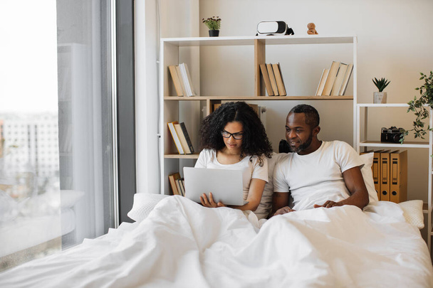 Beautiful young woman sitting in bed and sending text messages using modern laptop while her husband assisting her with advice. Loving married couple achieving tech-life balance resting at home. - Foto, afbeelding