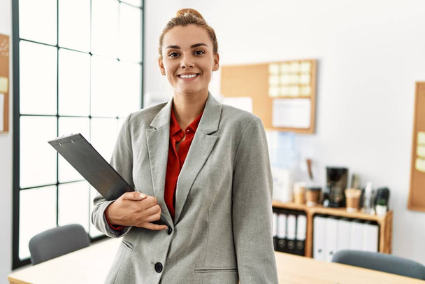 Νεαρή γυναίκα επιχειρηματίας χαμογελά αυτοπεποίθηση κρατώντας πρόχειρο στο γραφείο - Φωτογραφία, εικόνα
