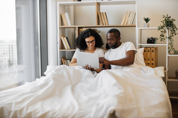 Beautiful young woman sitting in bed and sending text messages using modern laptop while her husband assisting her with advice. Loving married couple achieving tech-life balance resting at home. - Photo, image