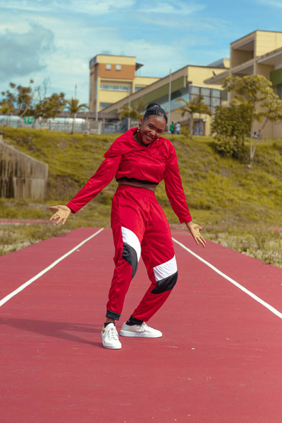 Mujer afro bailarina realiza baile en parque local en la pista de atletismo con hermosa vegetacion y cielo de fondo - 写真・画像
