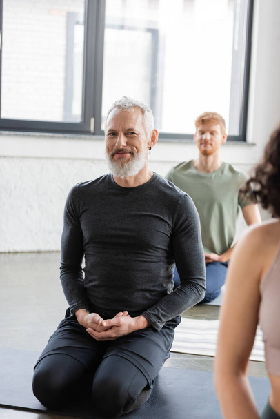 Smiling middle aged man looking at camera while practicing yoga in Thunderbolt asana in studio  - Foto, afbeelding