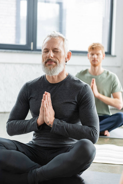 Uomo maturo meditando con gli occhi chiusi e anjali mudra sul tappeto in classe di yoga  - Foto, immagini