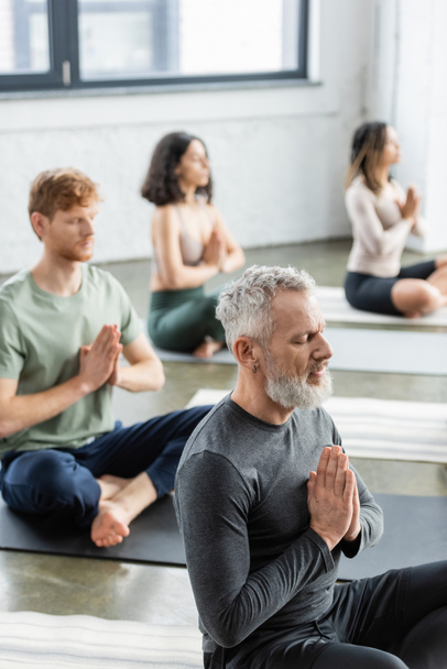 Mature man sitting in anjali mudra near blurred multiethnic group in yoga class  - 写真・画像