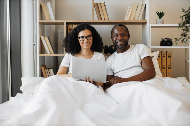 Intercultural romantic partners having rest under crisp white bedding during daytime with laptop in hands. Young modern man and woman forming healthy digital habits with balance to family at home. - Fotografie, Obrázek