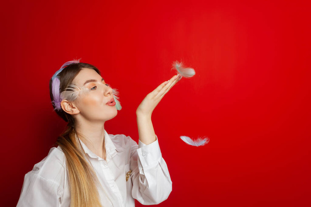 Portrait of a lovely tender girl wearing a feather decorations and blows away feathers from her hands. Isolated on a red background. - Foto, afbeelding