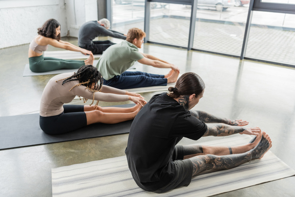 Side view of interracial people doing seated forward bend asana on mats in yoga class  - Foto, Imagen