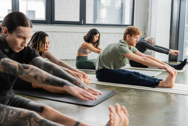 Young african american woman stretching on mat near people in yoga studio  - Photo, Image