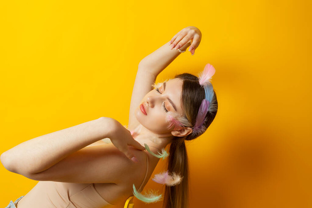 Portret van een lief teder meisje met veren decoraties en een korset topje dat zich bijna vermengt met de kleur van haar huid, poserend. Geïsoleerd op een gele achtergrond. - Foto, afbeelding