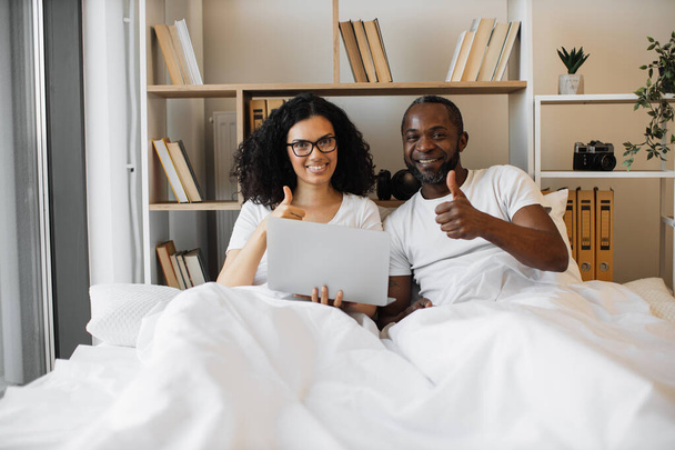 Χαρούμενο πολυφυλετικό ζευγάρι να κρατάει φορητό υπολογιστή και να σηκώνει τους αντίχειρες ενώ κείτεται κάτω από λευκή κουβέρτα στο σπίτι. Ρομαντικοί σύζυγοι που ενθαρρύνονται να ζουν άνετα. - Φωτογραφία, εικόνα