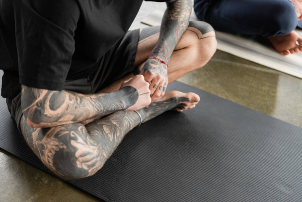 Vista recortada del hombre tatuado sentado en el tronco de fuego asana en la estera en la clase de yoga   - Foto, Imagen