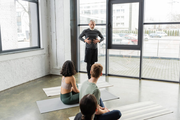 Entrenador de mediana edad hablando y haciendo gestos cerca de personas en esteras en clase de yoga  - Foto, Imagen