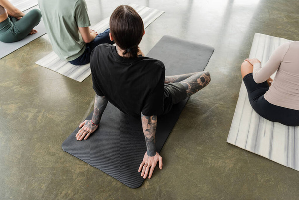 Vista aerea di uomo tatuato seduto su un tappeto vicino a persone interrazziali in classe di yoga  - Foto, immagini
