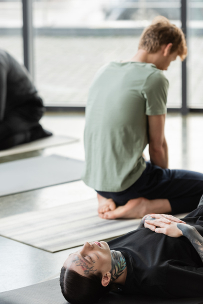 Getatoeëerde man ligt op mat in de buurt wazig mensen in yoga klasse  - Foto, afbeelding