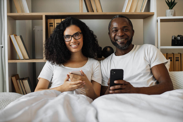 Serene multiracial family of two using mobile phones while lazing in soft bed during daytime at home. Confident adult man and bespectacled woman reading online article paying attention to details. - Zdjęcie, obraz