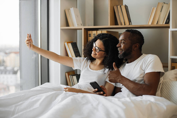 Cheerful african american spouses getting self-taken picture on smartphone while resting in studio apartment. Bearded man with phone in hand giving thumb-up sign while couple smiling at camera. - Fotografie, Obrázek