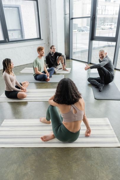 Lächelnder Trainer mittleren Alters im Gespräch mit einer gemischtrassigen Gruppe von Menschen auf Matten im Yoga-Kurs  - Foto, Bild