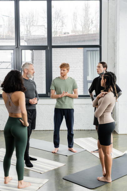Personas interraciales mirando al entrenador maduro mientras están de pie en las esteras en la clase de yoga  - Foto, Imagen