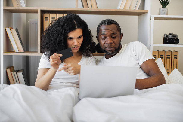 Young multiethnic spouses surfing internet via laptop while spending daytime at home. Smiling man and woman making reservation on e-commerce website using blank debit card without leaving bedroom. - 写真・画像